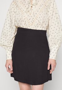 Saia preta justa com uma textura suave, apresentando um design simples. Combinada com uma blusa creme estampada com mangas bufantes.