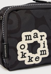 Μαύρο υφασμάτινο πουγκί με φερμουάρ, διακοσμημένο με ένα λευκό μπαλώμα σε σχήμα λουλουδιού που γράφει «marimekko» με μαύρα γράμματα.