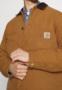 Hnědá plátěná pracovní bunda s tmavým límcem, dvěma náprsními kapsami a logem Carhartt na nášivce. Má zlaté kovové knoflíky a texturovanou tkaninu.