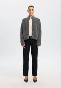 Suéter gris y peludo con cuello alto, delantero abierto y dos bolsillos en la parte delantera, combinado con jeans negros y tacones negros puntiagudos.