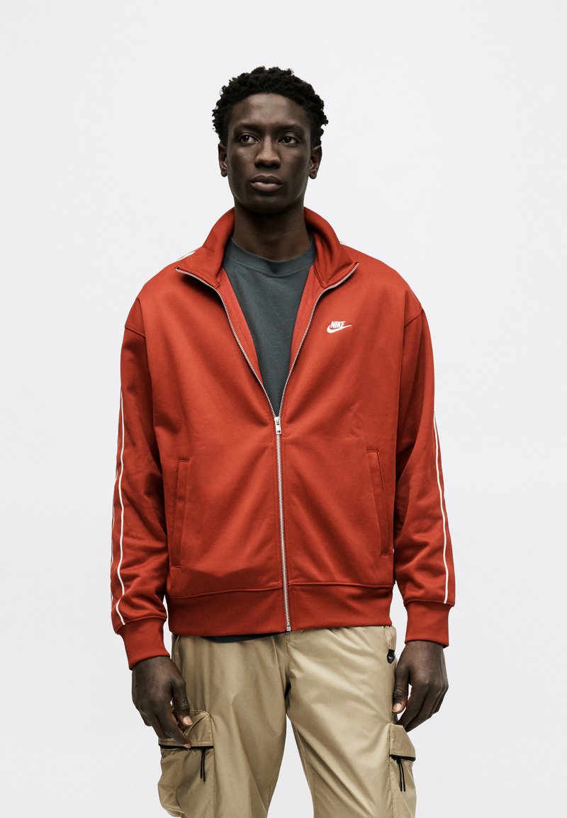 Uomo che indossa una giacca Nike rossa con cerniera sopra una maglietta grigio scuro e pantaloni cargo beige, in piedi davanti a uno sfondo chiaro e uniforme.