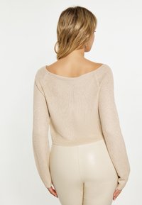 Beige lyhyhihainen neulepusero, jossa on pehmeä ja teksturoitu neule; siinä on leveä off-shoulder-kaula-aukko ja pitkät hihat, yhdistettynä istuviin kermaisiin housuihin.