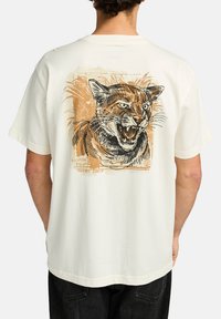 Camiseta color crema con un gran y detallado gráfico de un tigre rugiente en la parte posterior, con acentos en naranja y negro. Mangas cortas.
