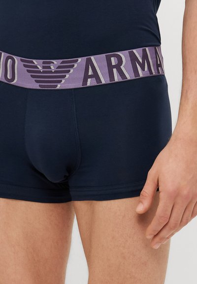 Tronco de algodão azul-marinho com um cós elástico roxo apresentando o logotipo "Armani" em cinza claro. Textura suave e design ajustado.