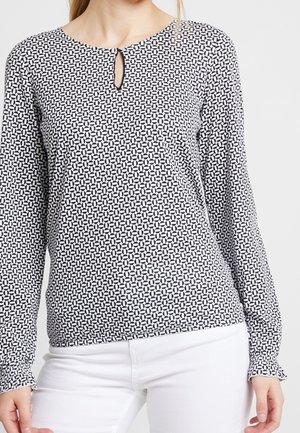 Žena v černo-bílé vzorované blůze s dlouhými rukávy a malým výstřihem ve tvaru klíčové dírky, doplněná bílými kalhotami.