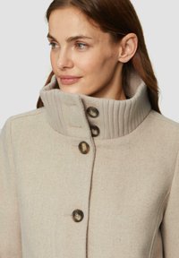 Beige wollen jas met een hoge geribbelde kraag, voorzien van drie grote bruine knopen aan de voorkant. Gladde textuur met subtiel verticale patroon.