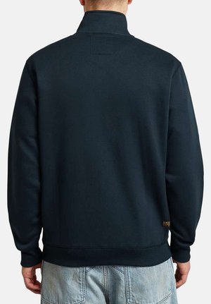 Persona che indossa una felpa a tinta unita blu navy scuro con maniche lunghe e colletto alto, vista da dietro, abbinata a un paio di jeans azzurri.