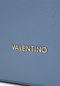 Blå strukturerad läder med en subtil kornmönster. Har guldpräglad "VALENTINO" text, som framhäver varumärkesidentitet och hantverk.