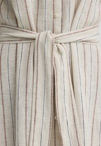 Svetlo bež tkanina z navpičnimi tankimi črnimi in rjavimi črtami, zavezana v ravno vozlje okoli pasu preko gumbov.