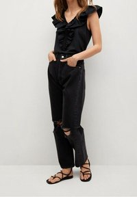 Débardeur noir à volants avec un col en V, associé à un jean noir déchiré taille haute et des sandales noires à brides.