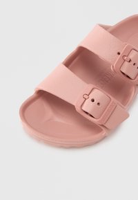 Lyserosa gummisandal med to justerbare remmer og spenner. Har en teksturert fotseng og en glatt såle med gripe mønster.