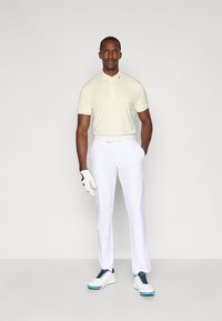 Lys beige polo-shirt, hvide bukser, hvide golf-sko med blå accenter og en hvid golf-handske. Modellen står foran en ensfarvet baggrund.