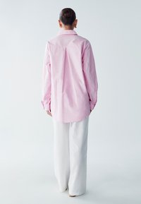 Camicia button-up rosa chiaro con piega sul retro, maniche lunghe e polsini, abbinata a pantaloni bianchi a gamba larga, indossata su uno sfondo neutro.
