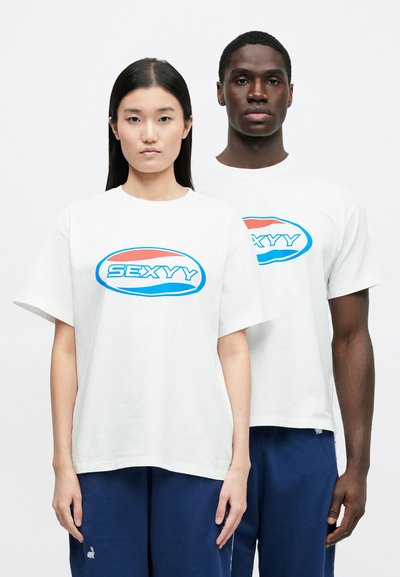 Vita bomulls-t-shirts med en blå och röd grafik med texten "SEXYY." Modeller bär marinblå joggingbyxor, visar korta ärmar och en avslappnad passform.