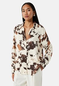 Beige overhemd met grote bruine en zwarte vlekken, voorzien van een knoopsluiting aan de voorkant en een relaxte pasvorm. Glad stoffen textuur.