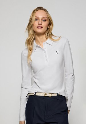 Polo shirt bianco a maniche lunghe con placket a bottoni e un piccolo logo nero. Indossato con pantaloni scuri e una cintura di colore chiaro. Texture liscia.