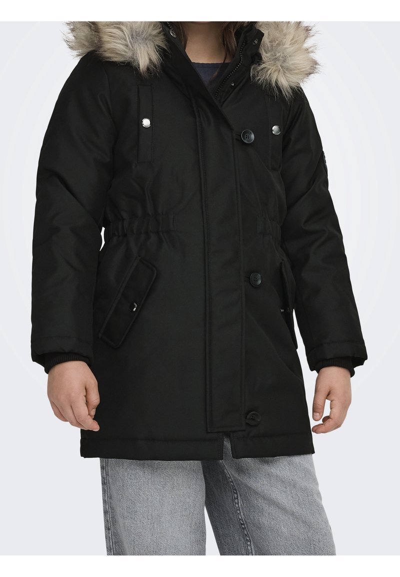 Enfant portant un manteau d'hiver noir avec capuche bordée de fourrure, fermeture à boutons et zip, taille élastique et jean gris clair.
