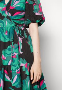 Vestido preto com um vibrante padrão floral em verde, rosa e roxo. As mangas bufantes, a cintura com laço e o design de saia em camadas são visíveis.