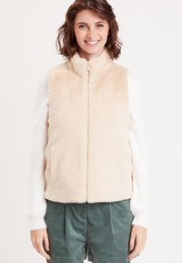 Gilet en fausse fourrure beige clair avec un col montant, fermeture éclair à l'avant et poches latérales, associé à un haut blanc à manches longues et un short vert.