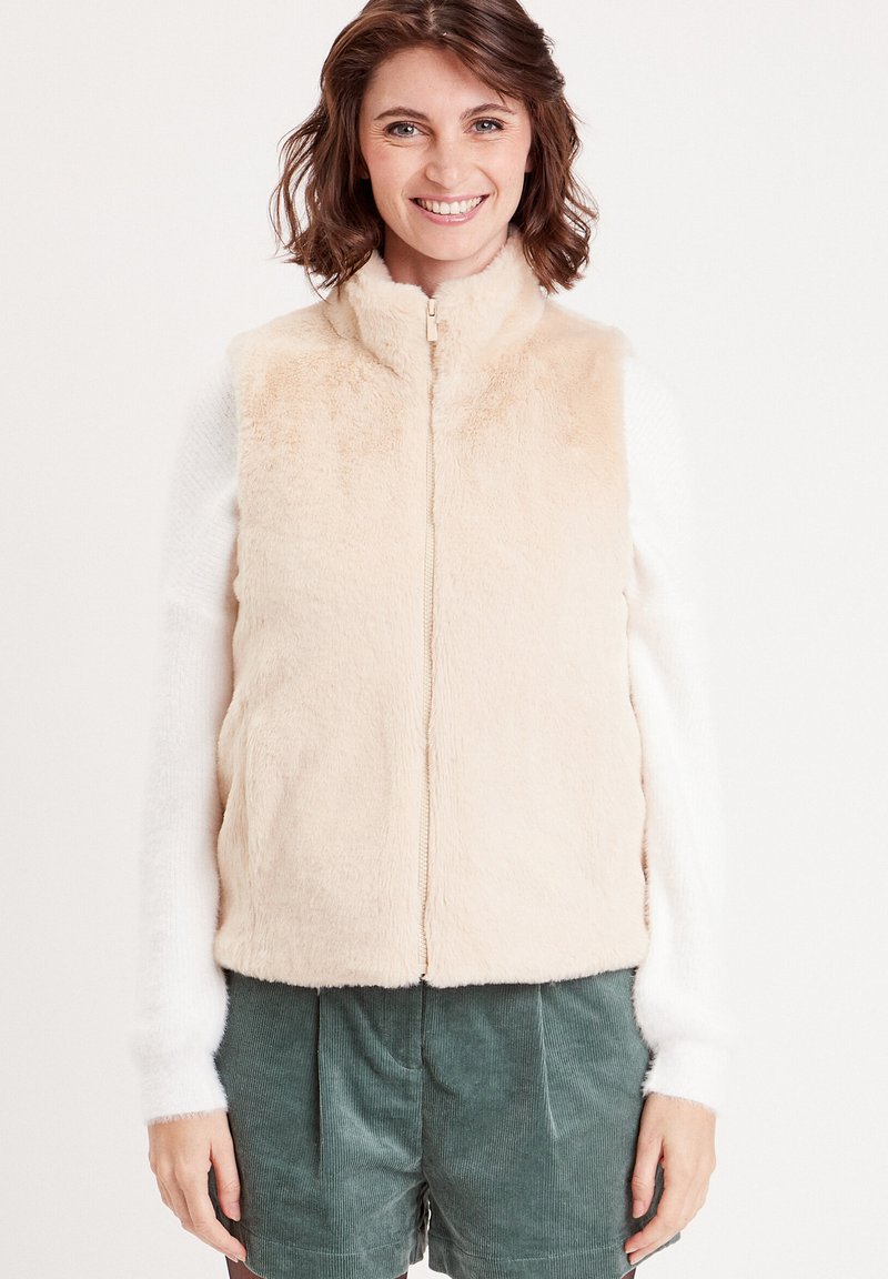 Gilet en fausse fourrure beige clair avec un col montant, fermeture éclair à l'avant et poches latérales, associé à un haut blanc à manches longues et un short vert.