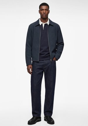 Mann stehend, trägt eine marineblaue Reißverschluss-Strickjacke über einem dunkelblauen Poloshirt, dunkle Jeans und schwarze Lederschuhe vor einem schlichten hellen Hintergrund.