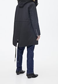 Czarna parka z kapturem i futrzanym obszyciem, wyposażona w rękawy na suwak i regulowane ściągacze u dołu, zestawiona z ciemnymi dżinsami i czarnymi butami.