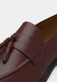 Καφέ δερμάτινο μοκασίνι με φούντες, λεπτομερείς ραφές και λείο, γυαλιστερό φινίρισμα στο πάνω μέρος του παπουτσιού.