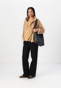 Tan fleece pullover with a high collar and zip closure, paired with black straight-leg pants and leopard print shoes, holding a black tote bag.