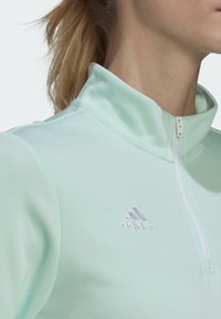 Türkisgrünes Sportoberteil mit einem Stehkragen, ausgestattet mit einem Viertelreißverschluss und einem weißen adidas-Logo auf der linken Brust. Glatte Textur.