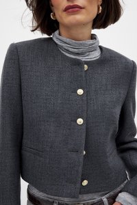 Harmaa teksturoitu takki, jossa on lyhyt muotoilu, korkealla kauluksella ja kullanvärisillä napeilla. Mukana etutasku ja kerroksellinen harmaa toppi alla.