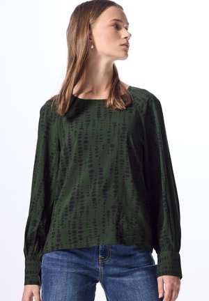 Blusa verde con motivo a pois neri irregolari, maniche lunghe e scollatura rotonda. Indossata con jeans blu, con polsini arricciati ai polsi.