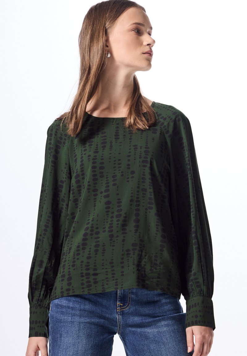 Blusa verde con patrón de puntos irregulares negros, de manga larga y cuello redondo. Llevada con jeans azules, con puños fruncidos en las muñecas.