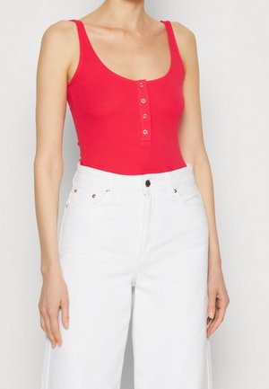 Una donna che indossa un top rosso senza maniche con bottoni e pantaloni bianchi a vita alta e larghi, in piedi contro uno sfondo semplice.