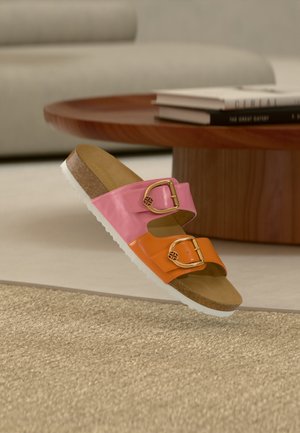 Sandalia tipo slide bicolor en rosa y naranja con hebillas doradas, plantilla de corcho y suela blanca, situada cerca de una mesa de café de madera y libros.