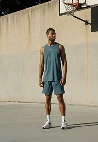 Άνδρας με μπλε-πράσινο αθλητικό φανελάκι και σορτς στέκεται σε εξωτερικό γήπεδο μπάσκετ κοντά στο καλάθι, κοιτάζοντας προς τα δεξιά.