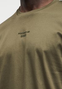 Κοντινό πλάνο ατόμου που φοράει ελιάς πράσινη μπλούζα με ανάγλυφο μαύρο λογότυπο «Porsche BOSS» στο στήθος, δείχνοντας τον λαιμό και το άνω κορμό.