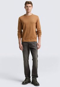 Męski sweter z okrągłym dekoltem w kolorze beżowym, zestawiony z ciemnoszarymi dżinsami i czarnymi butami sznurowanymi. Sweter ma prążkowane mankiety i dół.