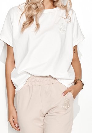 Vrouw die een wit T-shirt met korte mouwen en beige broek met elastische taille draagt, met een horloge aan haar linkerpols en stertattoos op haar rechterarm.