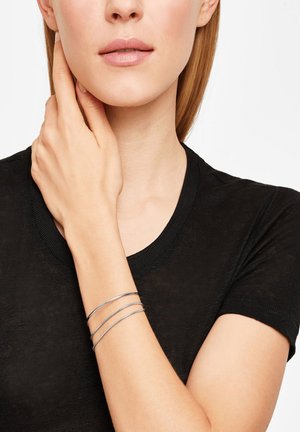 Zilveren manchetbands bracelet met drie dunne, verweven banden. Het model draagt het om haar pols, gecombineerd met een zwart aansluitend t-shirt. Glad textuur.