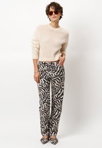 Strickcremepullover, gemusterte schwarz-beige Hose, gestreifte Schuhe und braune Sonnenbrille. Weicher Stoff und lockere Passform.