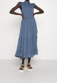 Robe maxi bleue à volants avec des manches froncées, décolleté en trou de serrure et taille élastique. Le modèle porte des sandales à plateforme. Tissu texturé avec des rayures verticales.