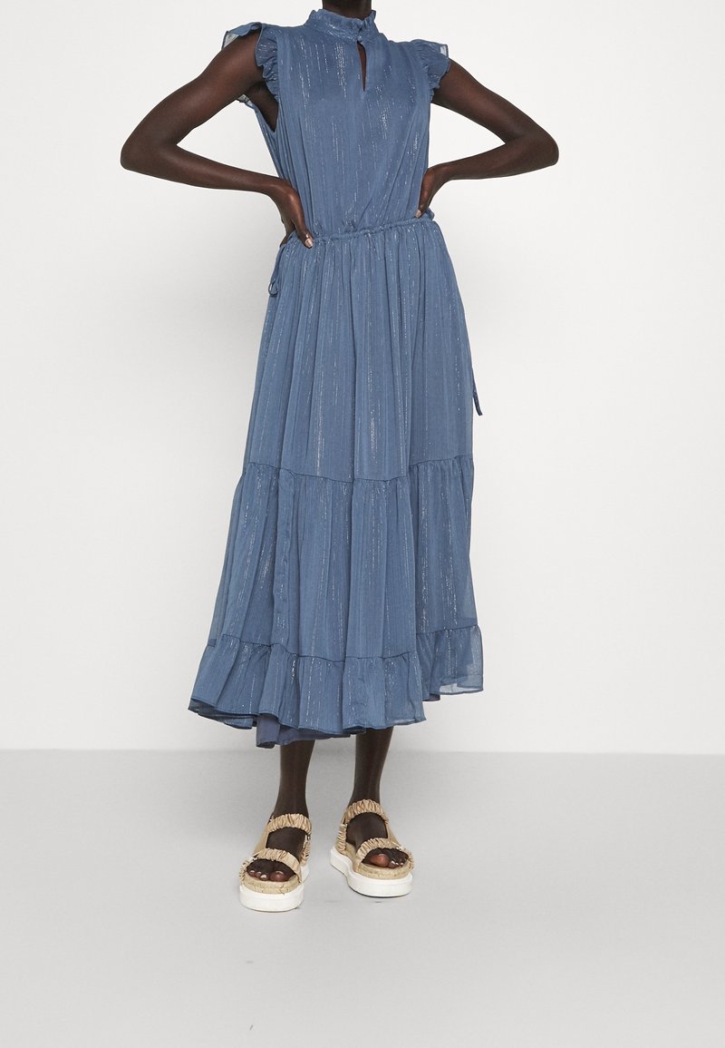 Robe maxi bleue à volants avec des manches froncées, décolleté en trou de serrure et taille élastique. Le modèle porte des sandales à plateforme. Tissu texturé avec des rayures verticales.