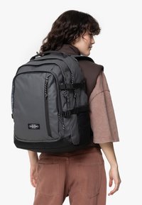 Zaino in tessuto grigio con accenti neri, dotato di numerosi scomparti, spallacci regolabili e dettagli con cordoncino.