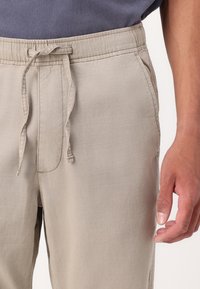 Lichtbeige broek met trekkoord, zachte textuur, elastische tailleband en zijzakken, vastgehouden door een hand tegen een donker shirt.
