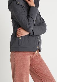 Szara ocieplana kurtka z kapturem, wyposażona w złote guziki, elastyczną talię i boczne kieszenie. Nosi się z różowymi sztruksowymi spodniami.