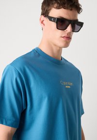 Kék pamut póló, kerek nyakkivágással és rövid ujjal, arany "Calvin Klein JEANS" logóval a mellkason. Matt fekete napszemüveg.