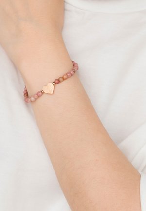 Arm mit einem Armband aus kleinen rosa Perlen und einem goldenen, herzförmigen Anhänger, der auf einem weißen Stoffhintergrund liegt.
