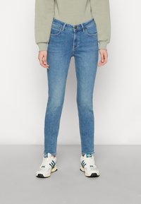 Persona con jeans ajustados de color azul claro, sudadera beige y zapatillas blancas con rayas verdes, de pie frente a un fondo liso.