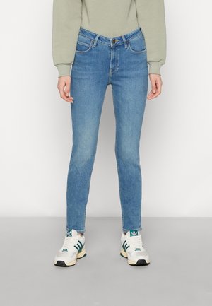 Persona con jeans ajustados de color azul claro, sudadera beige y zapatillas blancas con rayas verdes, de pie frente a un fondo liso.