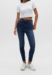 Mujer vistiendo jeans ajustados de cintura alta en azul oscuro, camiseta corta blanca y mocasines gruesos blancos, de pie contra un fondo liso.